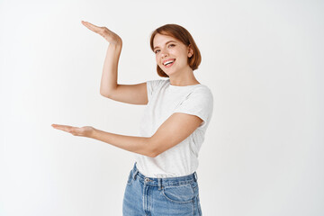 Beautiful caucasian girl with natural beauty face, showing big size object on copy space, shape large box with hands, standing on white background