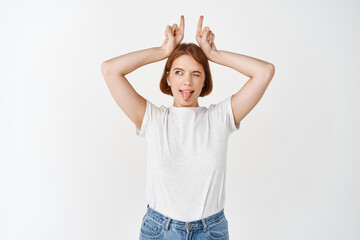 Happy young female model showing horns signs on head and tongue, looking aside with excited expression, standing on white background