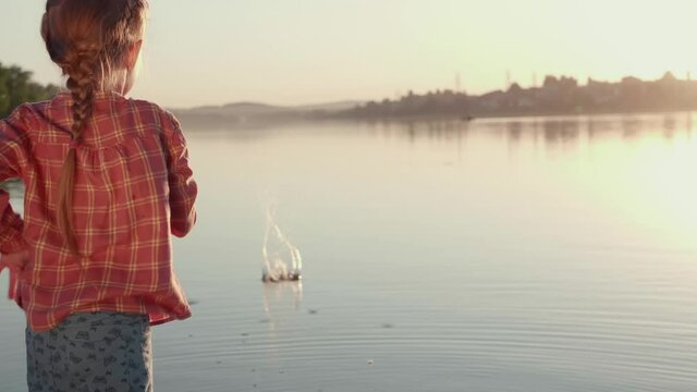 Back view on little girl in pink shirt playing with rocks.Child with blond pigtail throws pebbles in water on riverside, copy space.