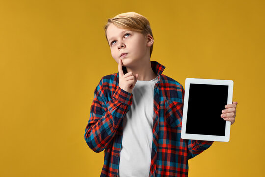 thoughtful child boy in checkered shirt shows empty tablet on yellow background