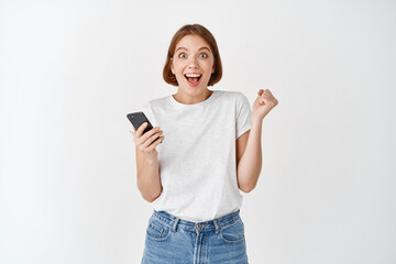 Fototapeta premium Happy woman winning on smartphone video game, raising hand up and cheering, shouting yes with joy, achieve online goal, standing on white background