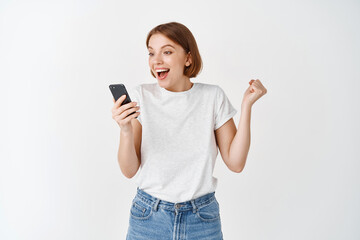 Fototapeta premium Happy woman winning on smartphone video game, raising hand up and cheering, shouting yes with joy, achieve online goal, standing on white background