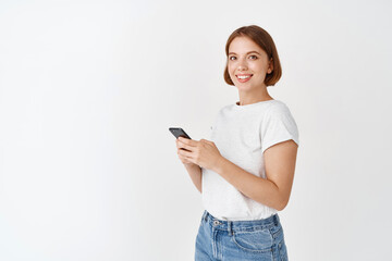 Fototapeta premium Young candid girl using smartphone and smiling at camera. Woman chatting on mobile phone app, standing against white background