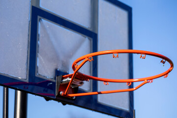 A portrait of an outdoor basketball rim without a net and a plexiglass backboard with blue lines on it in front of a blue sky. The basketball hoop or ring is orange and its pole is black.