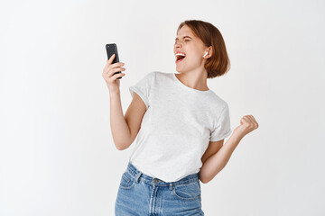 Fototapeta premium Portrait of carefree girl dancing with smartphone, listening music in headphones and smiling, standing on white background