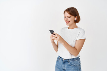 Young woman listen music in wireless headphones, looking at message on cell phone, reading screen and smiling, standing against white background