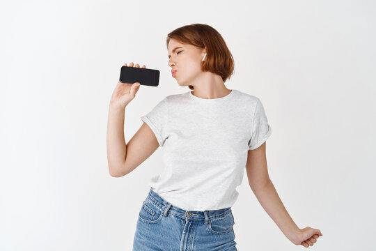 Carefree woman dancing and lipsync in smartphone, listening music in wireless headphones and showing phone screen, standing against white background