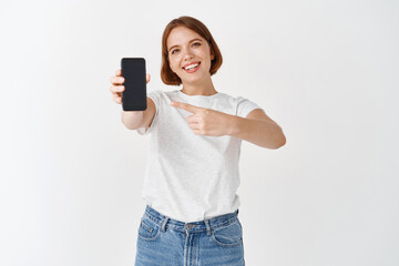 Check it out. Smiling beautiful woman in casual outfit, pointing at empty smartphone screen, showing online store or application, white background