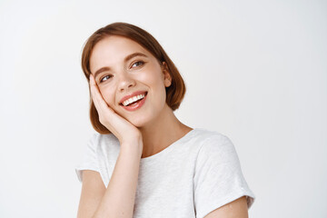 Fototapeta premium Beauty face. Tender happy woman with short hair, touching face and looking aside with dreamy smile, standing in t-shirt against white background