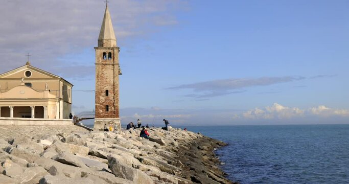 Church of the blessed virgin of the angel Madonna dell'angelo with people sitting on the stones on the sea shore with blue sky slow motion Caorle Italy