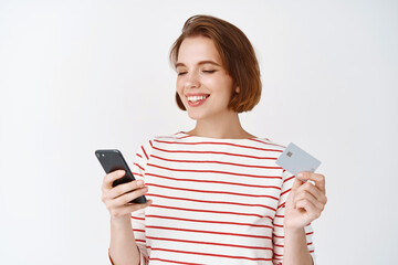 Young woman shopping online with plastic credit card and smartphone. girl reading mobile phone screen and smiling, paying in internet, white background
