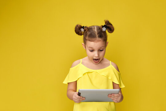 Surprised caucasian child girl in yellow t-shirt using digital tablet on yellow background