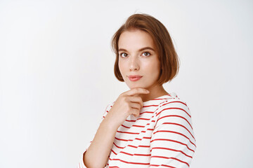 Obraz premium Portrait of thoughtful young woman looking intrigued at camera, touching face with natural, light make-up, standing on white background