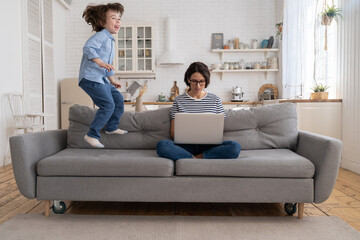 Focused young mother freelancer wear glasses sitting on couch working on laptop at home, trying to concentrate and not be distracted, hyperactive little son jumping on sofa and drawing attention. 