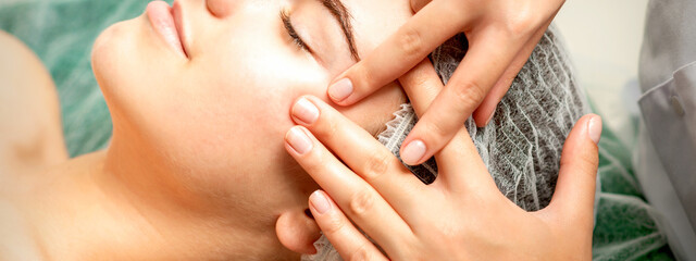 Young caucasian woman receiving facial massage by beautician's hands in spa medical salon