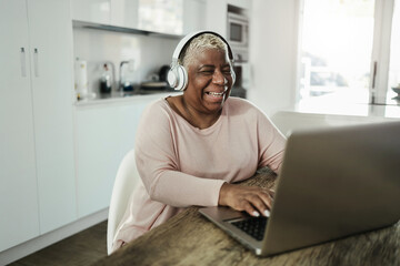 Senior woman using laptop while wearing headphones at home - Joyful elderly lifestyle and technology concept - Focus on face - Powered by Adobe