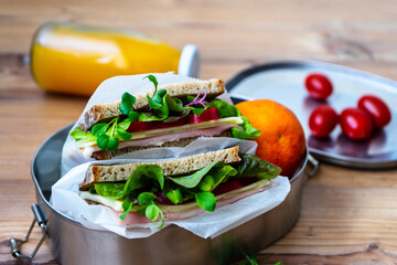 Zwei Sandwich mit Salat, Schinken und Käse in einer Blechdose auf einem Holztisch. Orangensaft in einer Glasflasche, Cocktail Tomaten, Nahaufnahme.