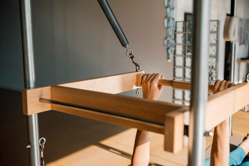 Hands detail of a woman doing Pilates, on a wooden Pilates Reformer machine, in a sunny gym indoors. Close up of a woman having a Pilates sports training.