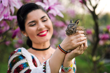 Beautiful young woman posing in romanian traditional costume with a little rabbit in a magnolia orchard