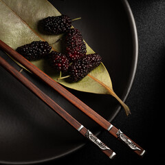 Black mulberries on green leaf and black dish with chopsticks in frame