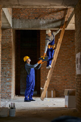 Help. Full length shot of young workman in blue overalls and hard hat giving toolbox to his colleague on ladder while working on house construction © Friends Stock