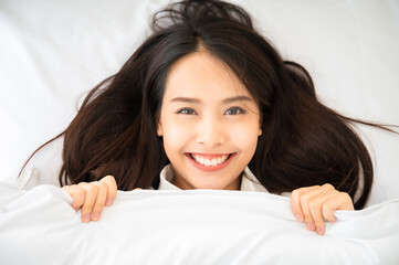 Young asian woman waking up in the morning on bed. She showing face out the blanket and smile...