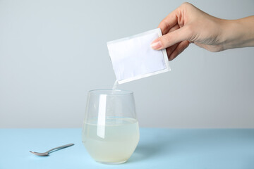 Woman pouring powder from medicine sachet into glass of water on turquoise table, closeup