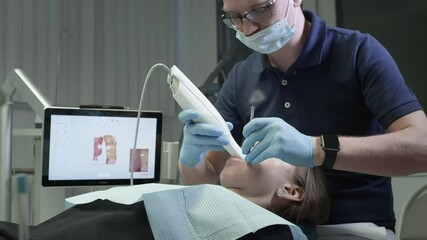 Doctor scans the patient's teeth in the clinic. The dentist holds in his hand a manual 3D scanner for the jaw and mouth. Dental health. Creates a 3D model of teeth and gums on a medical monitor. - Powered by Adobe