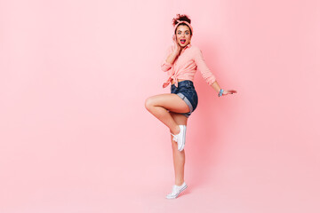 Fototapeta na wymiar Lady in pink shirt, denim shorts and sneakers jumps up and looks at camera with surprise