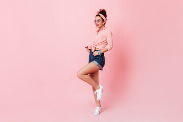 Fototapeta na wymiar Stylish pin-up girl with red lips lifts her leg on pink background. Full-length shot of woman in pink shirt and denim shorts