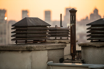 Chimney system on flat rooftop of residential building, with cityscape in background at sunset. Ventilation chimney with a rain cap and shielding. Smokestack pipes.