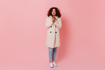 Full-length shot of young curly woman with red lips wearing white coat and jeans holding glass of tea on pink background