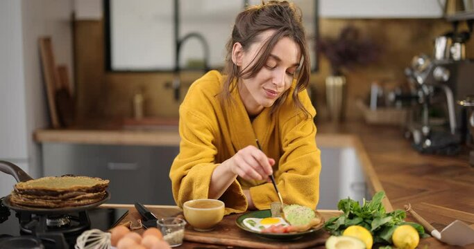 Young happy woman dressed in yellow bathrobe enjoyes healthy breakfast on the kitchen at home, eating avocado with fried eggs and greens