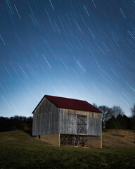 Obraz premium An old barn sits below a night sky as stars zip by in a long exposure.