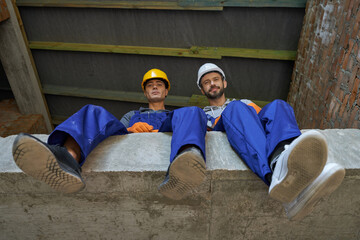 Two attractive young builders in blue overalls and hard hats looking down at camera, sitting on the...