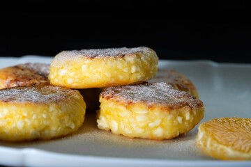 Sweet cottage cheese pancakes with orange slices on a white plate close-up against a black wall with copy space.