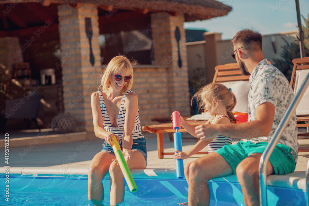 Wall mural Parents playing with their daughter by the swimming pool, splashing water on each other