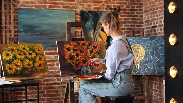 Young beautiful female artist painting still life with sunflower on canvas using oil paintings and palette knife. Painter creating artwork in art studio. Relaxation, leisure, hobby, stress management.