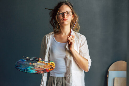 Artist female posing in her art studio. A professional candid young woman painter in transparent eyeglasses searching for an imagination in the workshop.
