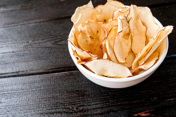 Chips of fresh apples with cinnamon are on a white plate on a background of black wooden boards. Organic apple chips. Dried fruits. Healthy sweet snack. Dehydrated and raw food. Copy space.