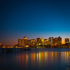 Fototapeta na wymiar Boston Nightscape Skyline and Water Light Reflections on Mystic River. The Modern City of Boston and Weathered Damaged Dock Pilings. Harmony of Nature, Civilization, and Passage of Time.