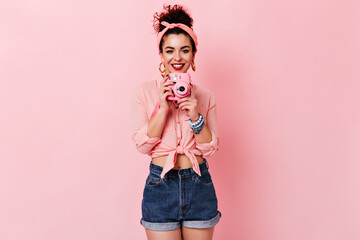 Curly lady in pink blouse and denim shorts is smiling and holding mini camera on isolated background
