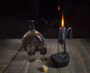 An old dusty bottle of dark brown bottle with alcohol, two goblet and a metal candlestick holder with a candle on a wooden table on a dark background.