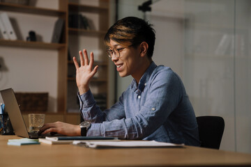 Smiling asian guy waving hand while making video call on laptop