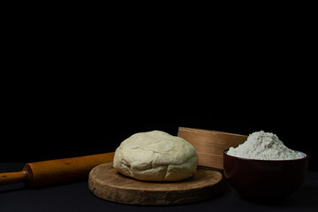 Dough on a dark background. A piece of homemade dough on a wooden board on a black background. The tools for working with the baking dough lie around the dough.