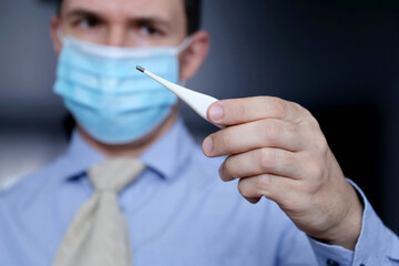Worried man in face mask and office clothes looks at digital thermometer in hand. High body...