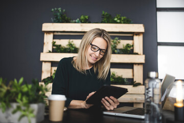 pretty young woman with glasses sits in a modern, sustainable office with lots of green ecological plants and works on her tablet and is happy and is smiling