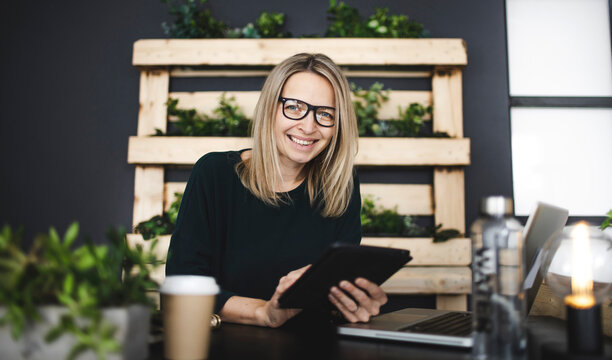 Pretty Young Woman With Glasses Sits In A Modern, Sustainable Office With Lots Of Green Ecological Plants And Works On Her Tablet And Is Happy And Is Smiling