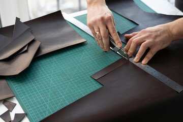 Men's hand holding a stationery knife and metal ruler and cutting on a pieces for a leather wallet...
