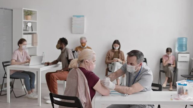 Medium long of male Caucasian doctor wearing mask and gloves, sitting at table, giving vaccine shot to blonde senior woman in busy hospital room, patients waiting on background